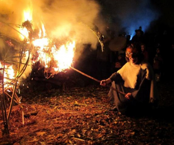 A man in Iron Age costume and wearing a mask lights a wicker bonfire,
