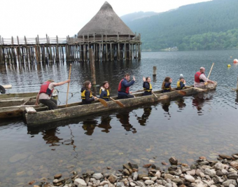 A team of eight people paddle a wooden long boat out onto Loch Tay with the Crannog Centre in the background.