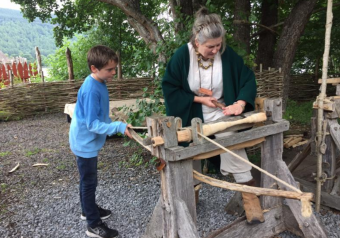 A woman in Iron Age costume works a piece of wood on a frame, while a child watches.