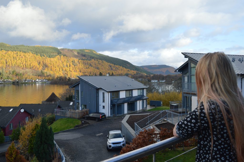 Photo of the view from a balcony overlooking an accommodation complex with loch and valley beyond.