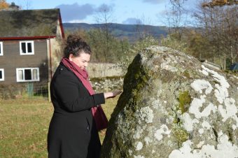 Photo of a woman gingerly touching a standing stone in a field.