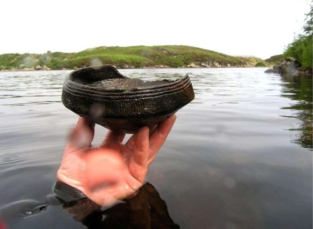 Photo of a hand holding a broken bronze bowl out of a loch