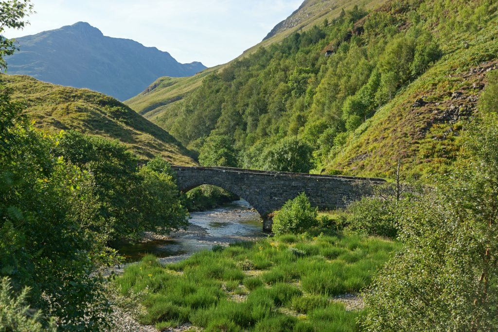 Photo of a valley floor with a river running along the bottom and a grey stone arched bridge set over the river.