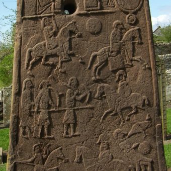Photo of a large standing stone slab with cravings depicting soldiers and men on horseback engaged in battle.