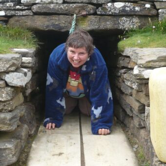 A woman with short brown hair and a blue fleece, crawling through a prehistoric stone corridor with a wide smile on her face.