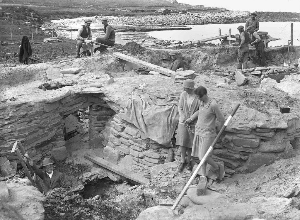 Black and white photo of two women observing a dig trench in a prehistoric settlement.