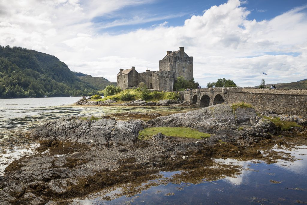 Photo of a small square medieval-like castle on an island in a loch, connected to the mailand by a small arched bridge.