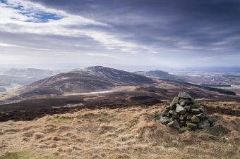 Photo of a low Scottish peak in the distance, covered in light brown heather and gorse, under a purple sky.