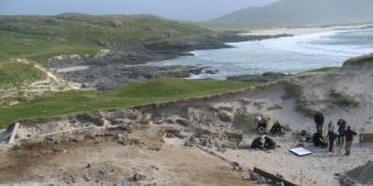 Photo of a Scottish beach, with the sea in the background and a film crew and excavation team on the sand dunes in the foreground.