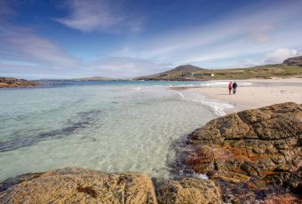 Photo of a couple walking along a white sandy beach with blue skies overhead.