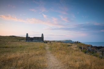 Photo of a small ruined church building on a cliff by the sea with a cal, blue evening sky.