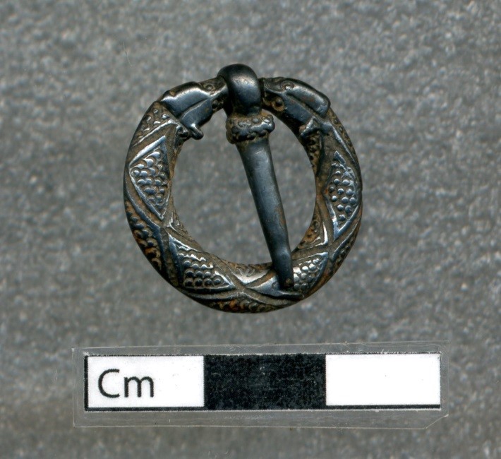 Photo of a small ring-shaped, metal brooch and pin with the shape of two dog heads.
