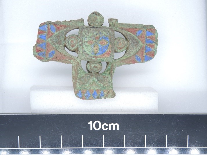 Photo of a tiny cross-shaped badge for a horse harness, with the top arm missing, and the remains of blue and red paint decoration on the front.