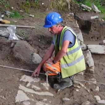 Archaeologist bending down near a trench pointing to something