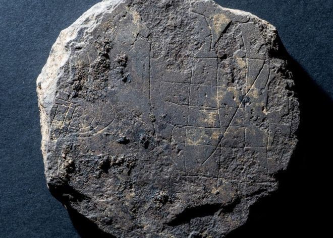 Photo of a round, flat piece of grey stone, with faint etchings of a game board on top.