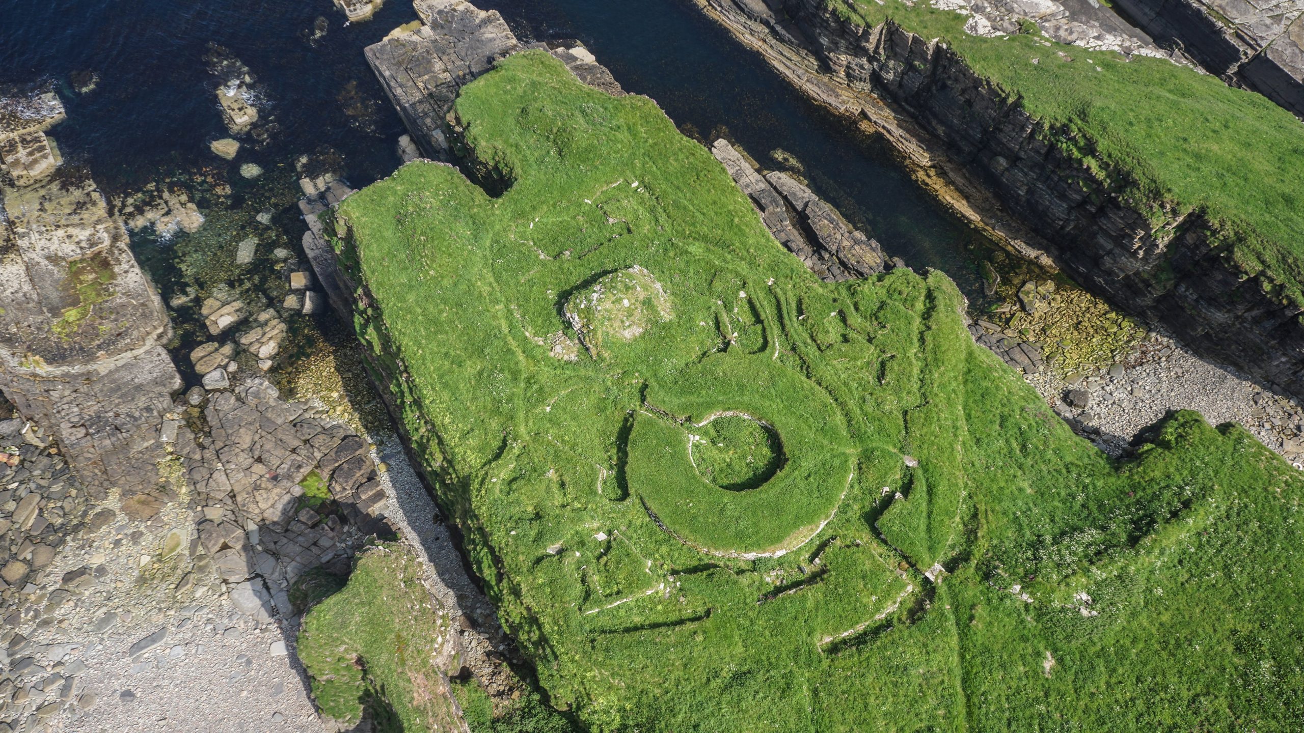 Aerial photo of earthwork remains from a prehistoric stone roundhouse by the sea.