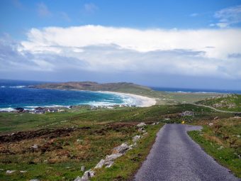 Photo of a winding tarmac road with the sea to the left and clouds overhead.