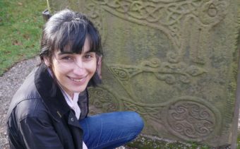 Photo of a young person kneeling by a carved stone slab and smiling at the camera.