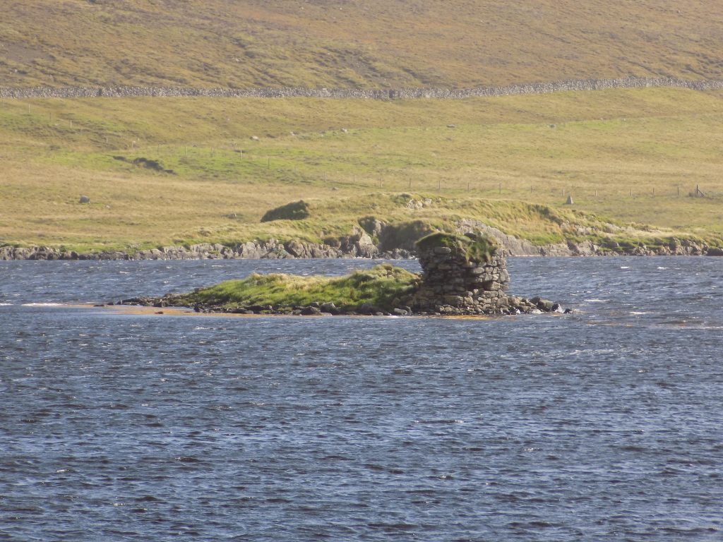 Photo of a collapsing low stone wall, the last remains of a building on a small islet in the middle of a loch.