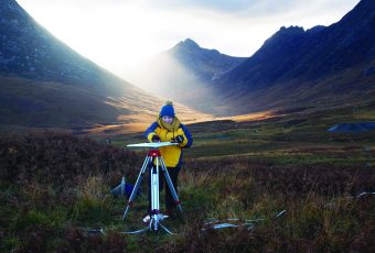 Photo of a person in a big coat and hat drawing on a board held on a tripod in the middle of a huge valley.