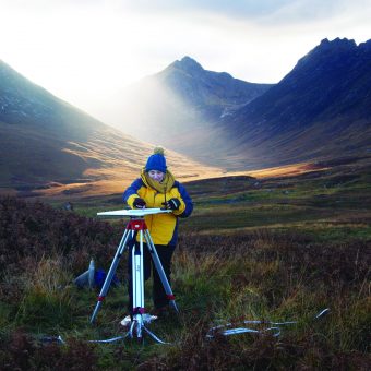 Photo of a person in a big coat and hat drawing on a board held on a tripod in the middle of a huge valley.
