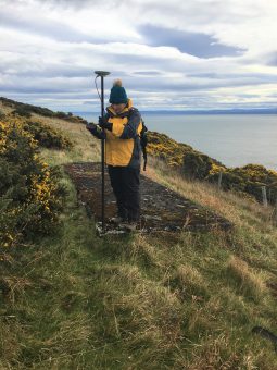 Photo of a person in a big coat and hat holding an instrument on a pole and taking electronic readings from it, by the sea.