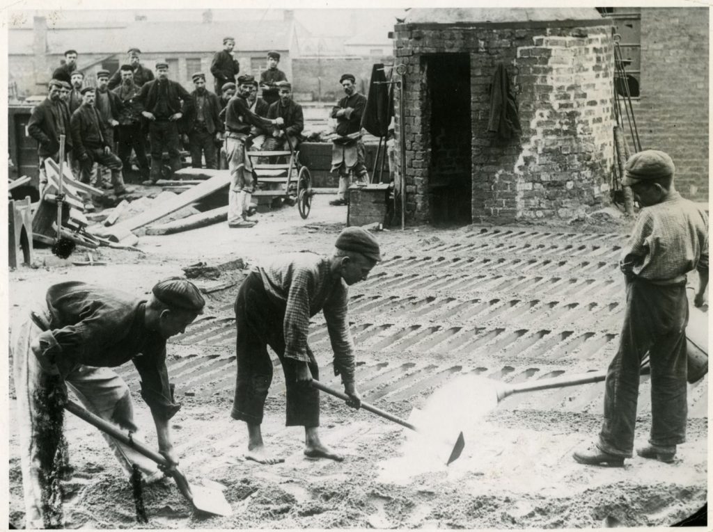 [IMAGE 6: Boys preparing pig iron moulds in a bed of casting sand at Coltness Iron Works, Newmains around 1910. In the background the men of the furnace team wait to ‘tap’ the blast furnace.]