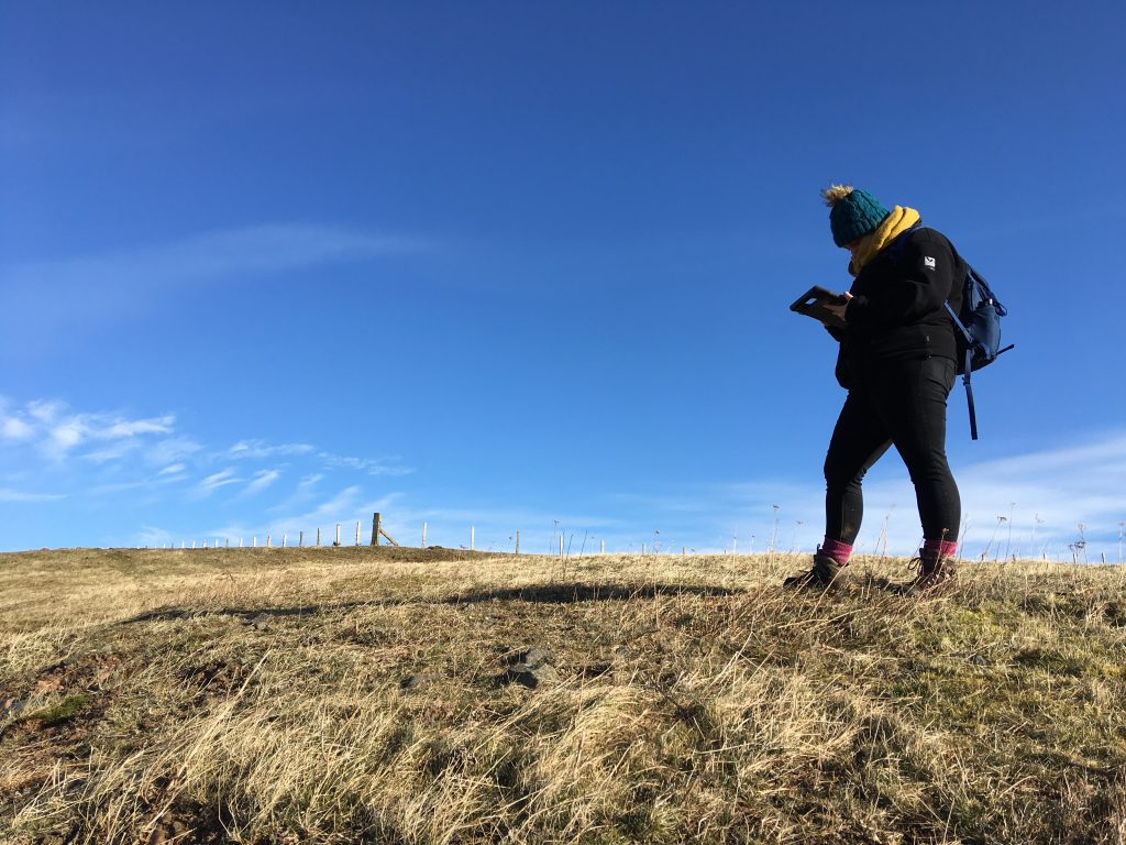 Photo of a person in a big coat and hat looking down at a tablet while walking through a field.