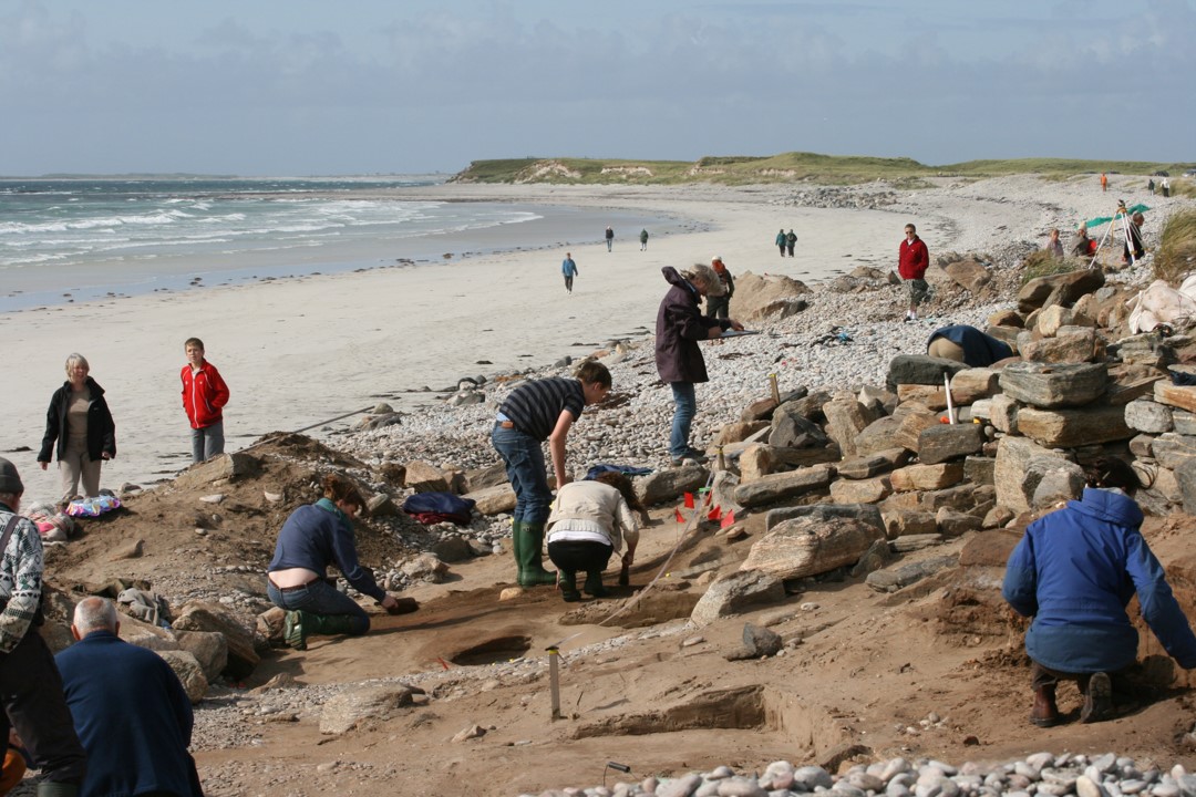 Photo of a large group of people working to look through an archaeological site on a beach close to the water.