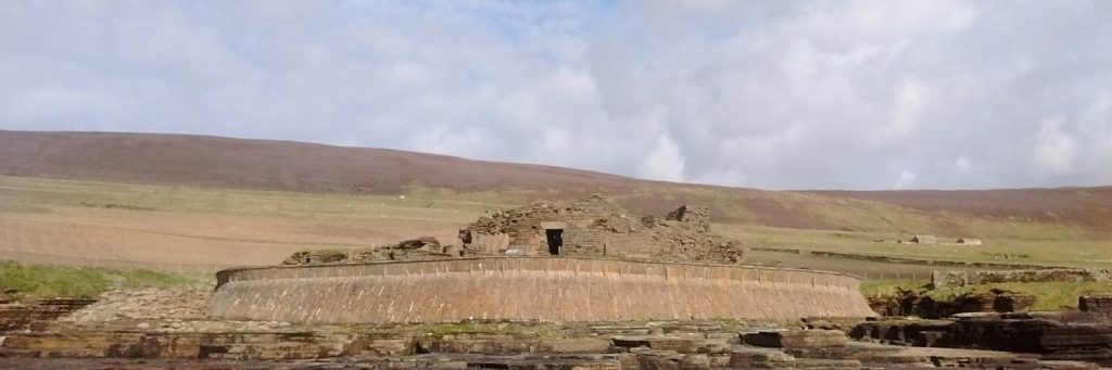Photo showing a ruined prehistoric circular stone dwelling, surrounded by a modern sea wall defence.