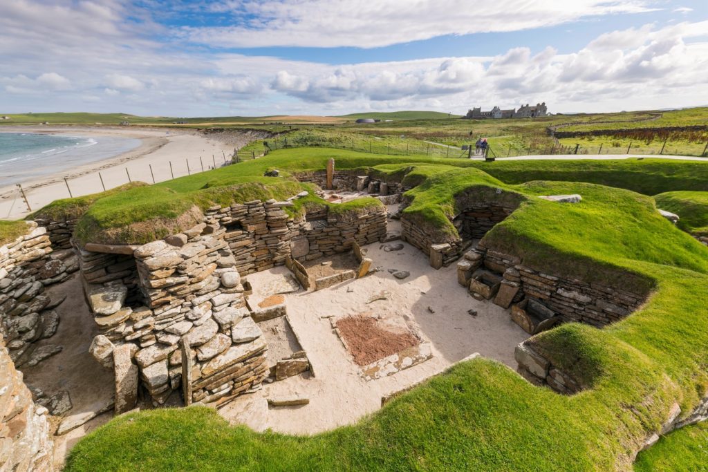 Photo of the remains of a prehistoric village beneath grassy dunes, next to the sea.
