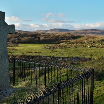 Celtic style cross surrounded by a small, black metal fence, overlooking green hills