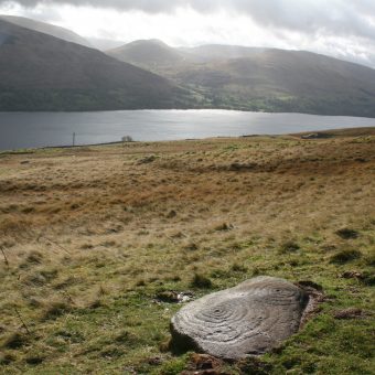 Photo of a large flat stone in a field by a loch with mountains in the background.