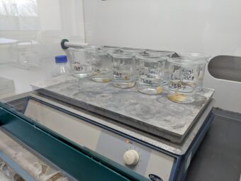 Photo of samples of human bone in beakers of clear liquid being heated on a metal plate.