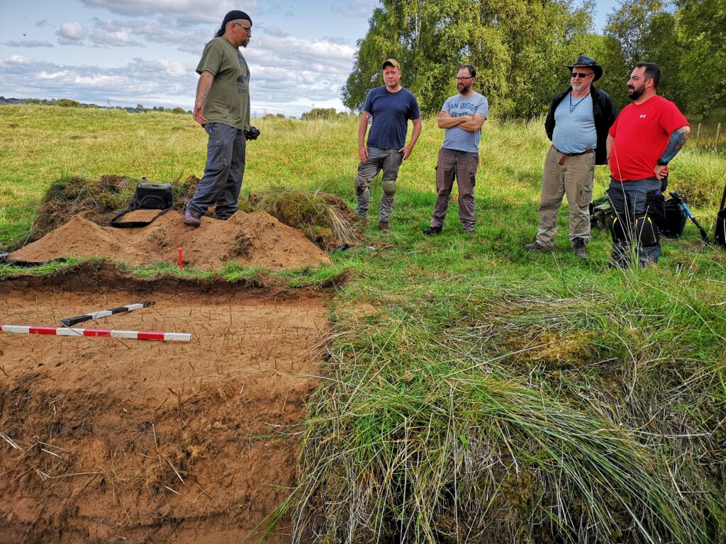 Photo of five people standing at the side of an archaeological trench in a field