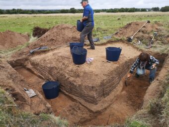 Photo of the corner of an archaeological trench with three black buckets and a person in view.