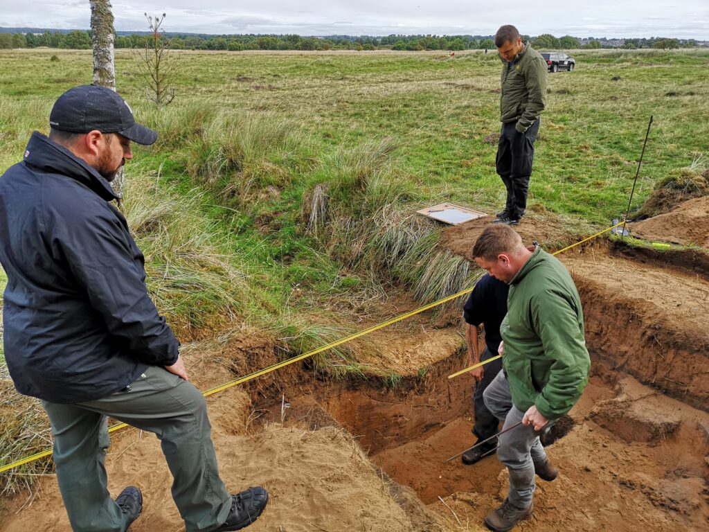 Photo of three people in an archaeological trench in a field measuring the site.