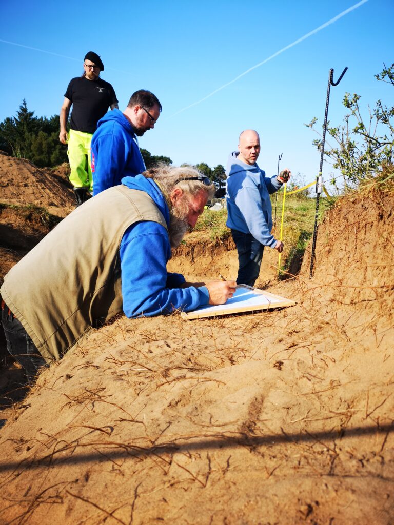 Photo of a man leaning on the side of an archaeological trench making notes on paper.