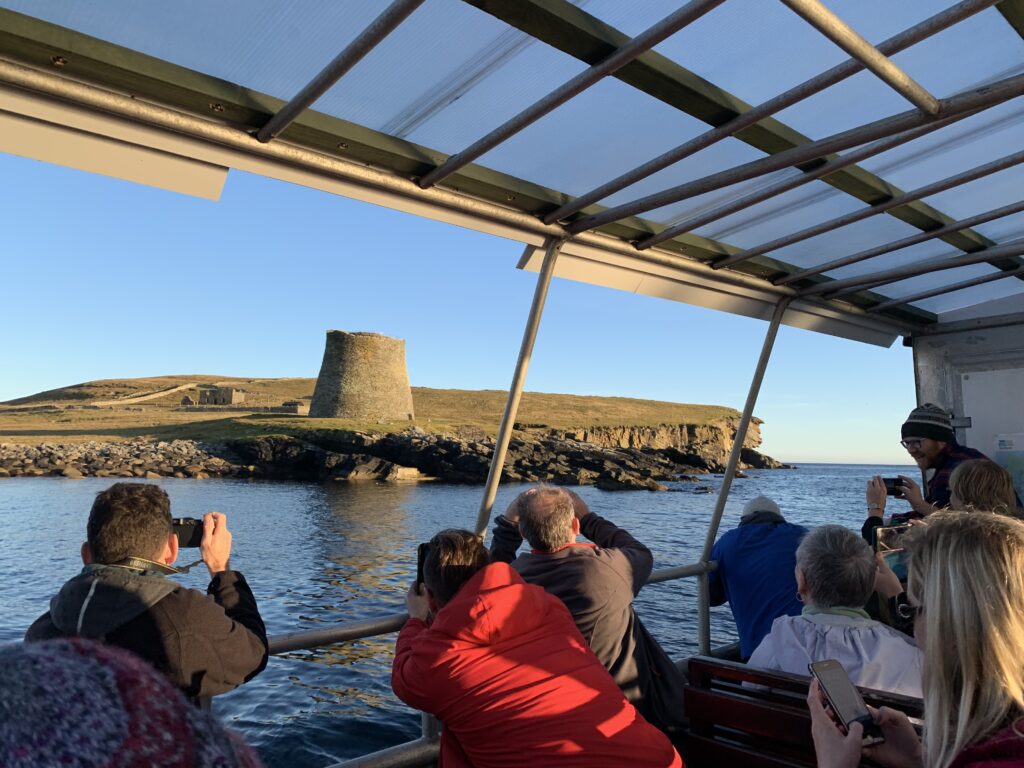 Photo of people on a boat taking pictures of an ancient round tower across the water on an island