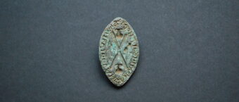 Photo of a small, greenish metal shield-shaped seal matrix with a visible St Andrew's cross shape etched on the face