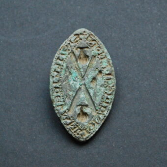 Photo of a small, greenish metal shield-shaped seal matrix with a visible St Andrew's cross shape etched on the face