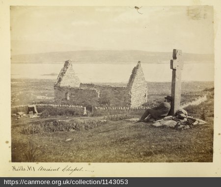 Early photo of a small, ruined medieval chapel on an island with the sea in the background, and a standing stone cross in the foreground