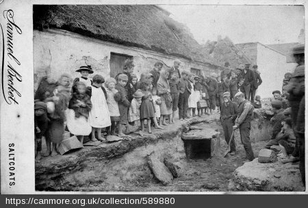 Early black and white photo of a rural street dug up to reveal a stone, square cist (ancient container), with children lining the street to see it