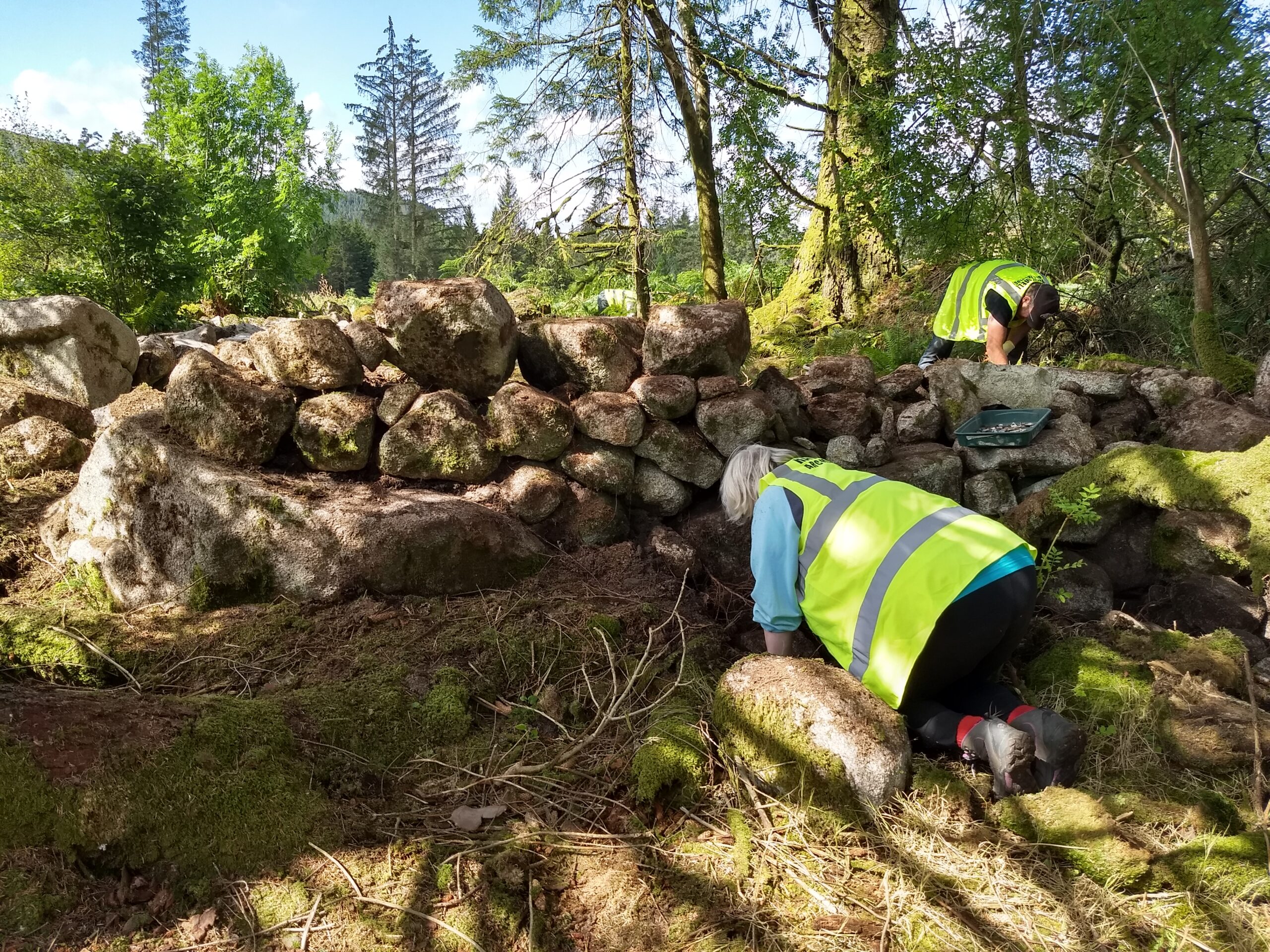 Photo of two people in high-vis jackets kneeling by a wall in a forest