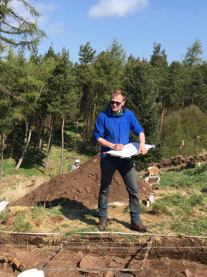Photo of a person in sunglasses and a blue fleece standing next to an archaeological trench and drawing on a small whiteboard