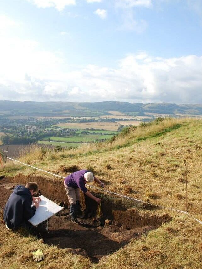 Photo of a person digging in an archaeological trench while another person sits and draws