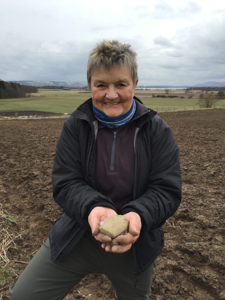Photo of a person in a muddy field holding out a small stone and smiling to the camera