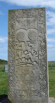 Photo of a grey, carved standing stone slab featuring a battle scene and symbols and knots