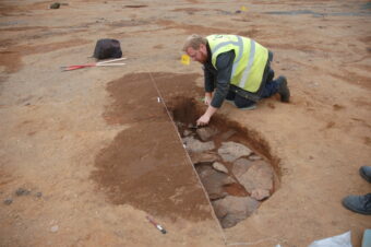 Photo of a person in a reflective vest kneeling by an open archaeological trench