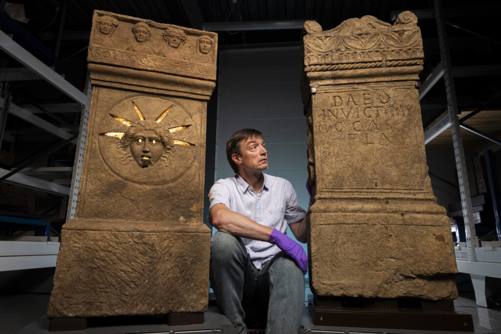 Photo of a man kneeling between two stone Roman altars